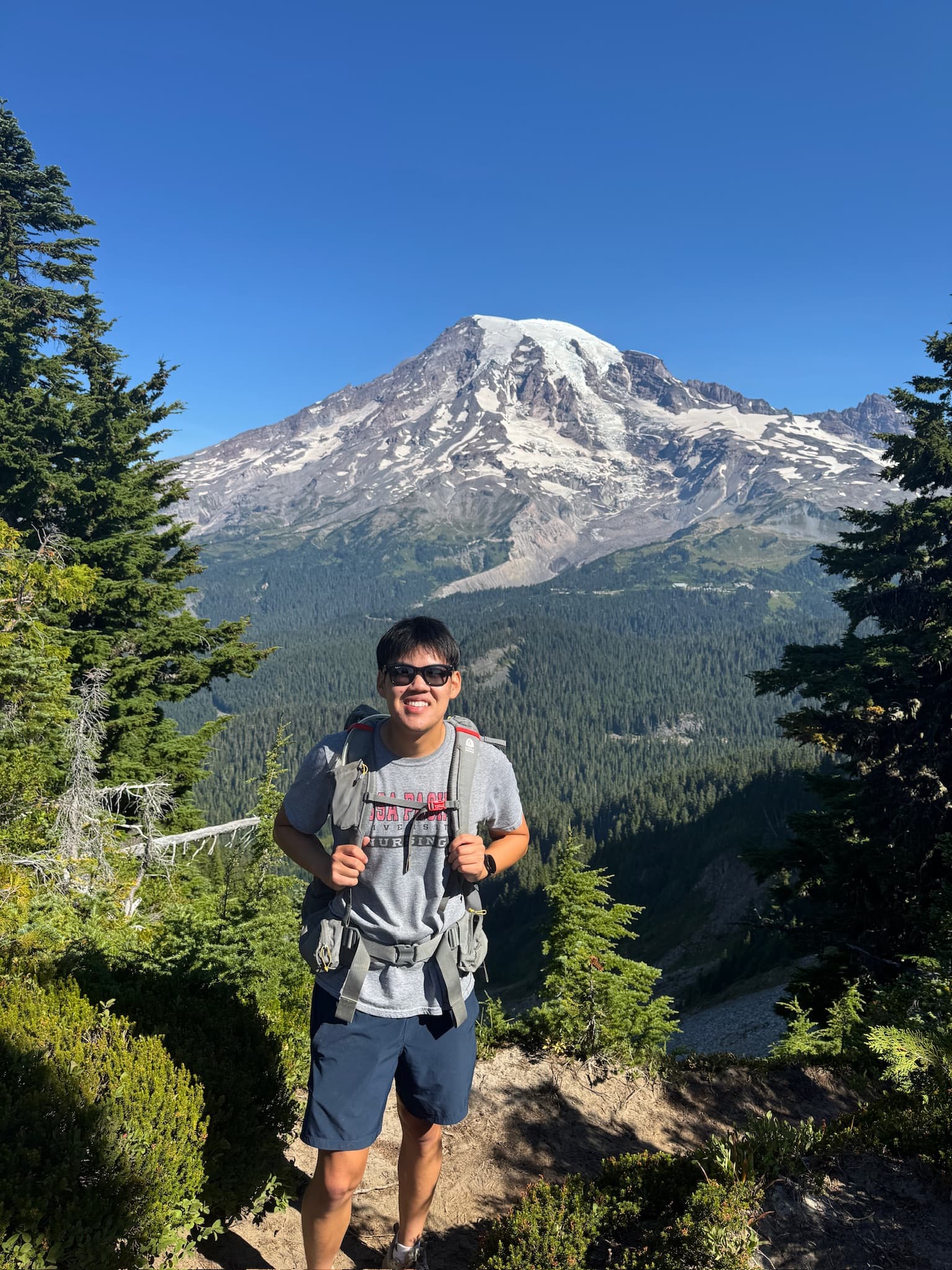 Jethro Chu wearing a backpack on a mountain trail, with forest and a snow-covered peak behind him.